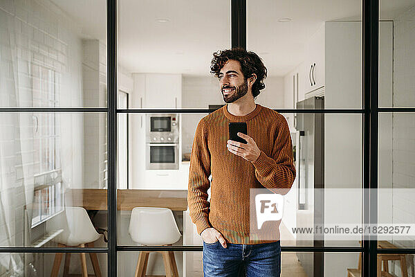 Glücklicher junger Mann mit Smartphone  der zu Hause an einer Glastür lehnt