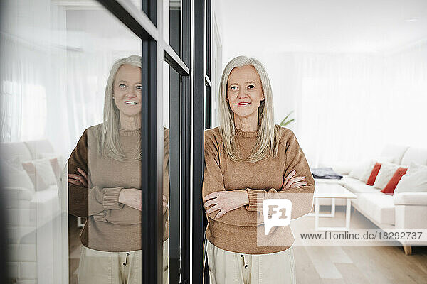 Smiling mature woman with arms crossed standing in living room at home
