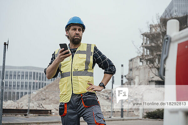 Worker holding smart phone with hand on hip in front of construction site