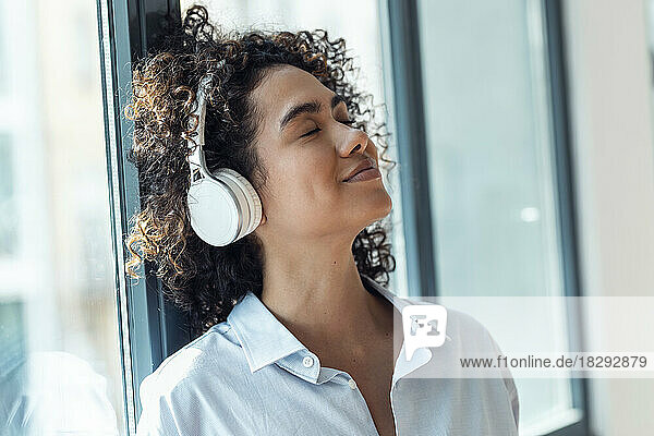 Smiling woman wearing wireless headphones leaning on window at home