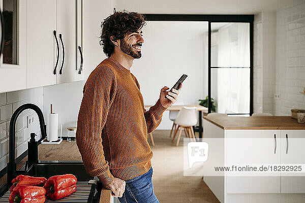 Happy young man standing with mobile phone by kitchen counter at home