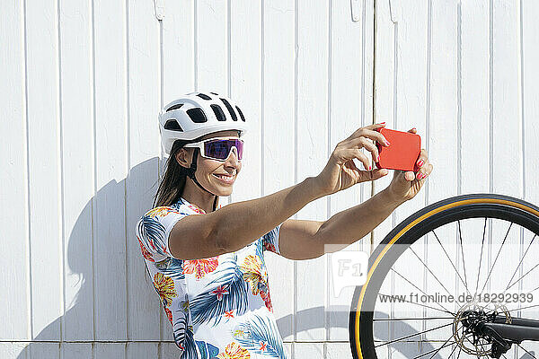 Happy cyclist taking selfie through smart phone in front of wall