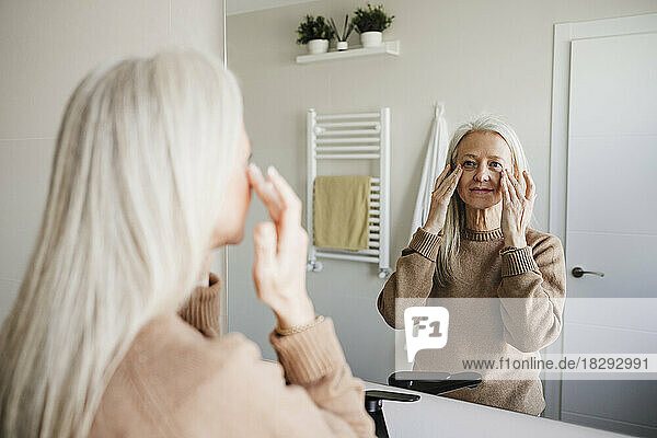 Mature woman looking in mirror and applying moisturizer on face