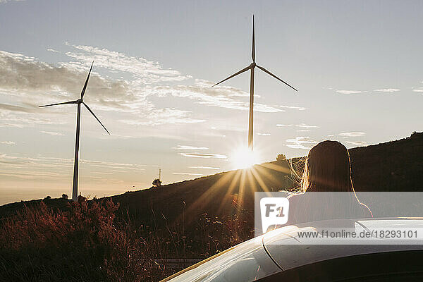 Woman leaning on car looking at wind park at sunset