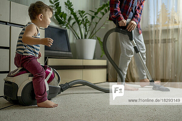 Boy cleaning carpet with brother sitting on vacuum cleaner at home