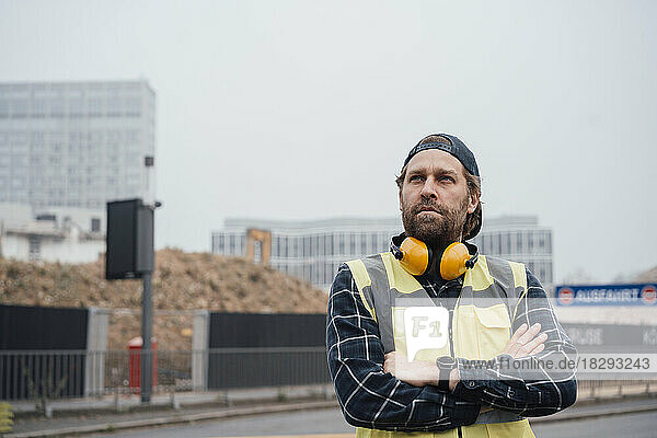 Thoughtful mature worker with arms crossed standing in front of construction site