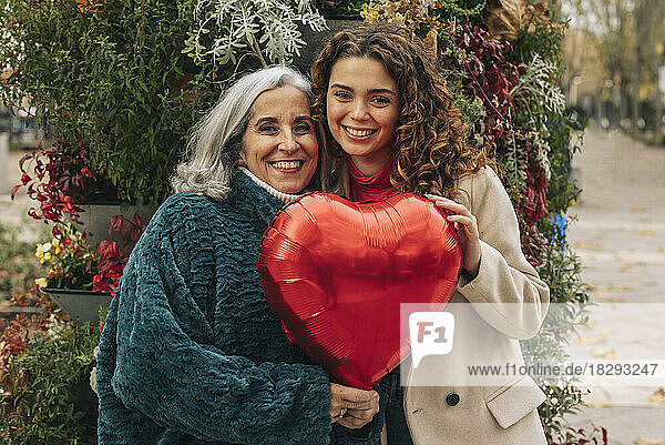 Happy granddaughter and grandmother holding red heart shaped balloon