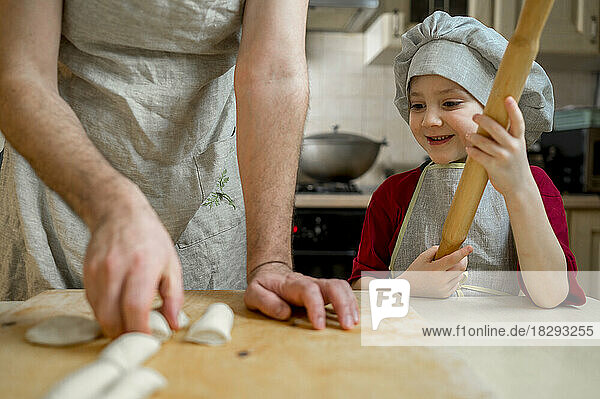 Smiling boy with rolling pin looking at father preparing dough on kitchen counter