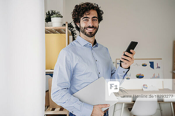 Smiling young engineer holding smartphone and laptop in office