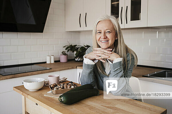 Happy mature woman with hands on chin sitting at table in kitchen