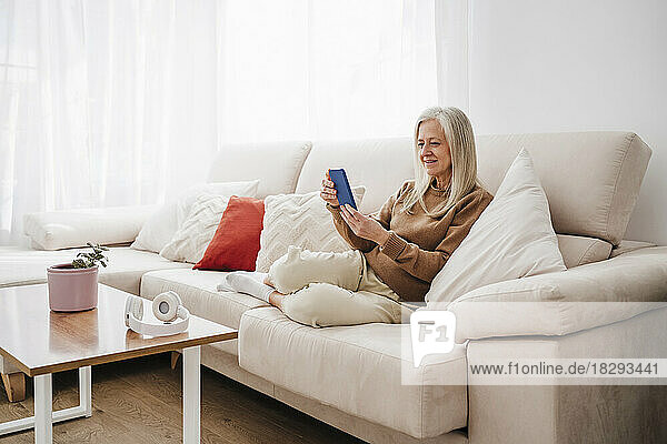 Lächelnde reife Frau  die zu Hause auf dem Sofa sitzt und ihr Smartphone benutzt
