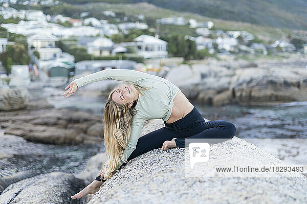 Smiling young woman doing stretching exercise on rock by sea