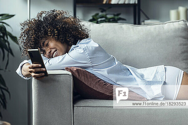 Smiling woman using smart phone lying on sofa at home