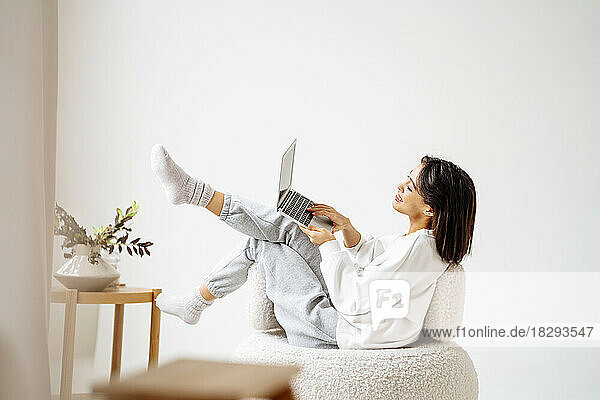 Woman using laptop sitting on chair by wall at home