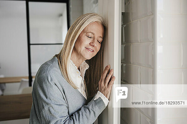 Smiling mature woman leaning on radiator with eyes closed at home