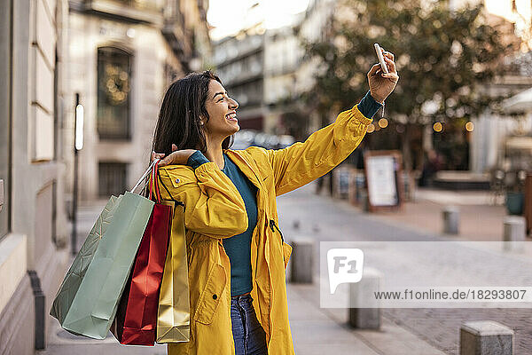 Happy woman with shopping bags taking selfie over smart phone on footpath