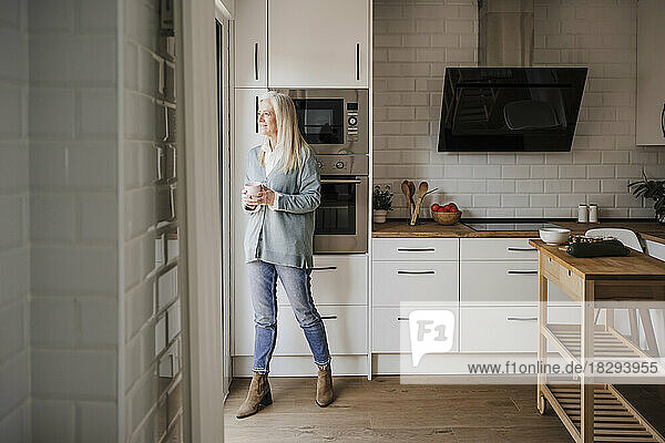 Nachdenkliche reife Frau steht mit Kaffeetasse in der Küche