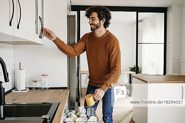 Happy young man with cup opening cabinet in kitchen at home