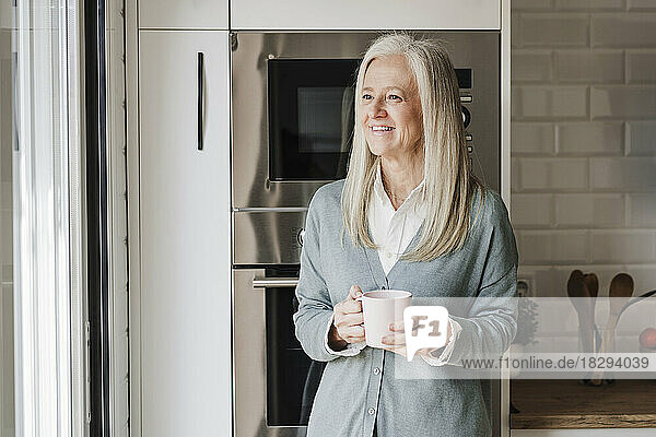 Happy mature woman with coffee cup standing in kitchen