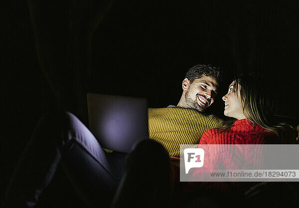 Happy couple using laptop at night smiling at each other