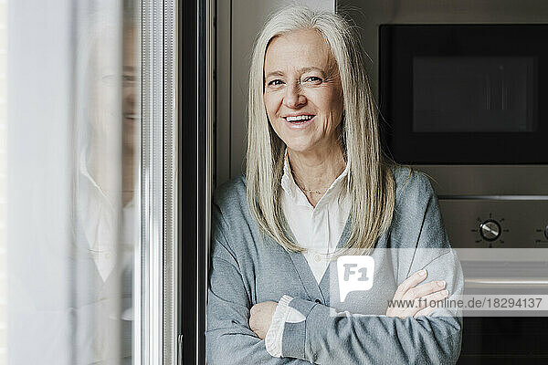 Happy mature woman with arms crossed standing by window at home