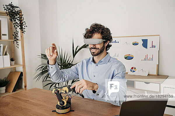 Happy businessman wearing virtual reality glasses with robotic arm on desk at office