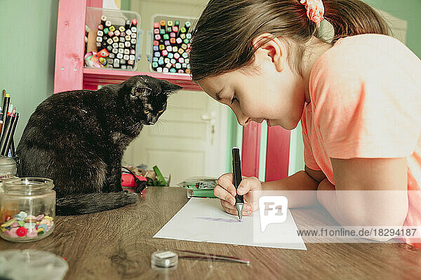 Katze beobachtet Mädchen  wie sie zu Hause ein Bild auf Papier zeichnet