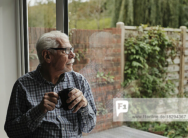 Nachdenklicher älterer Mann mit einer Tasse Kaffee  der zu Hause aus dem Fenster schaut