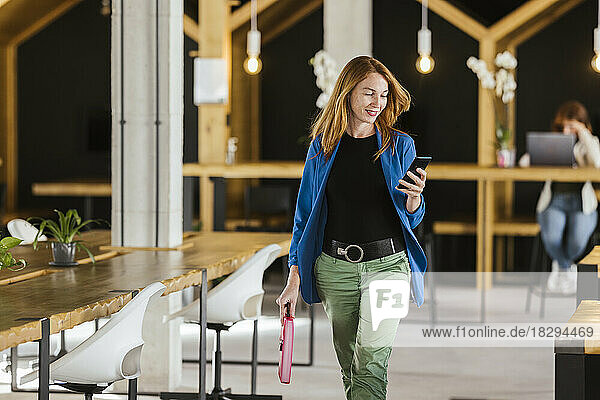 Happy businesswoman walking and using smart phone in office