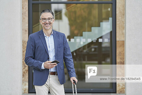 Smiling businessman holding smart phone standing in front of building