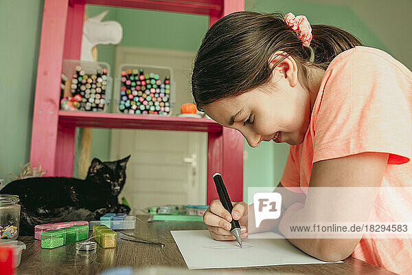 Lächelndes Mädchen  das auf Papier zeichnet  während die Katze am Tisch sitzt