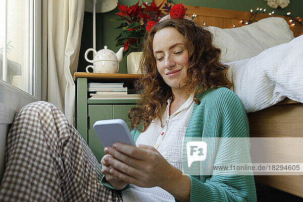 Smiling woman using smart phone sitting by bed at home