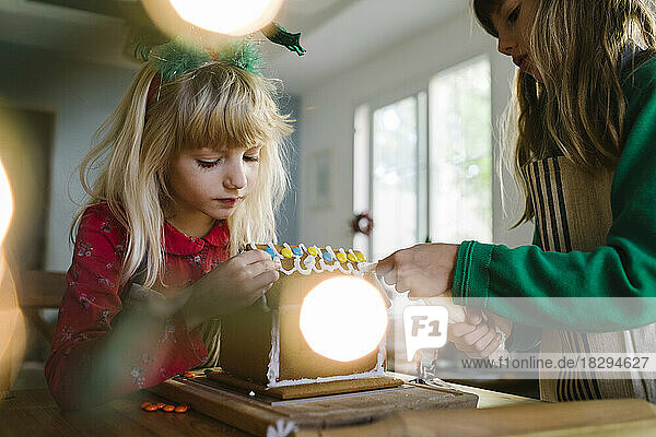 Sisters decorating gingerbread house with cream at home