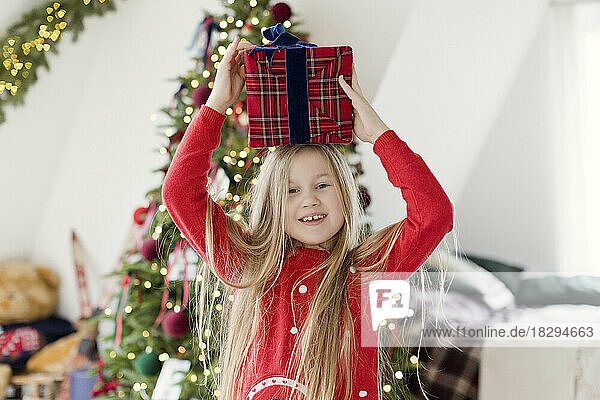 Fröhliches blondes Mädchen hält zu Hause ein Weihnachtsgeschenk auf dem Kopf