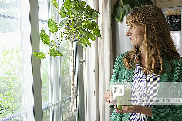 Smiling woman with green smoothie standing by window at home