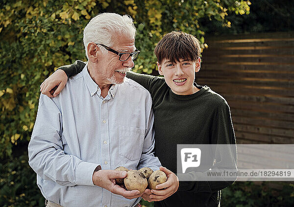 Happy grandfather and grandson standing in garden holding freshly hervested poatoes