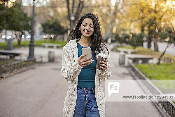 Happy woman with disposable cup using smart phone in park