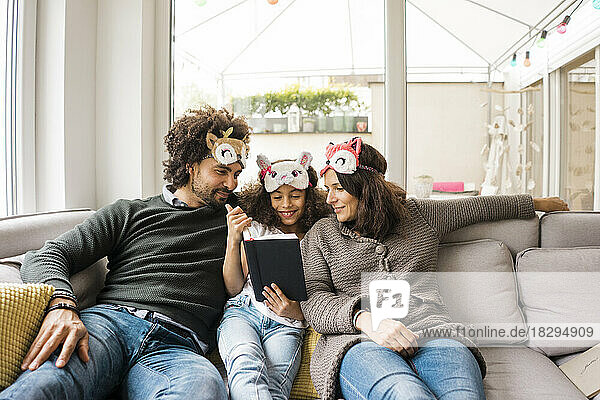 Lächelnde Eltern mit Tochter  die Augenmasken trägt und zu Hause auf dem Sofa ein Buch liest