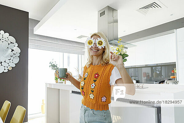Smiling woman wearing flower sunglasses standing with tea cup in kitchen at home