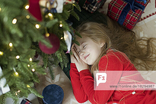 Smiling blond girl relaxing by Christmas tree on rug at home