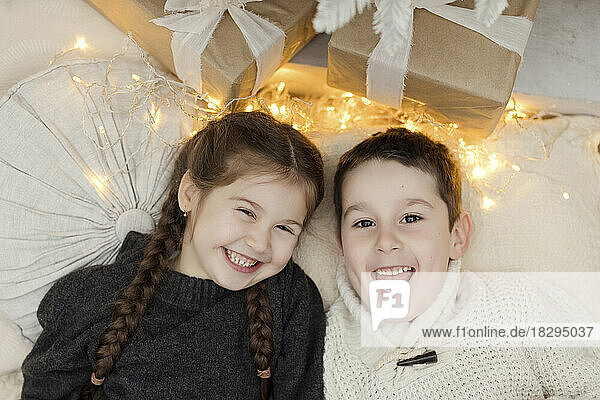 Happy girl and boy lying by Christmas lights and gifts at home