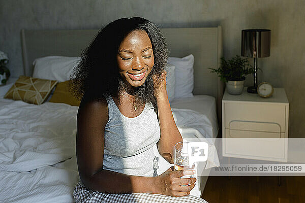 Happy woman with eyes closed holding drinking glass on bed at home