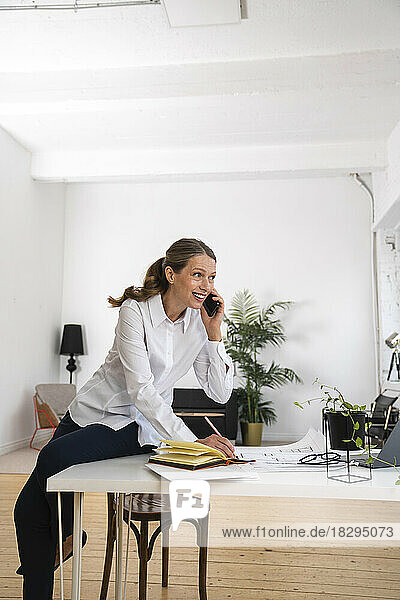 Glückliche reife Geschäftsfrau  die am Schreibtisch im Büro sitzt und über ihr Mobiltelefon spricht