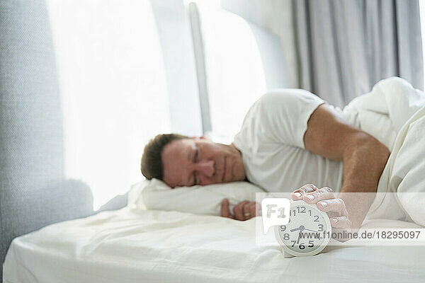 Man holding alarm clock lying on bed in bedroom