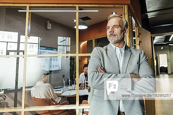 Thoughtful mature businessman with arms crossed leaning on glass wall in office