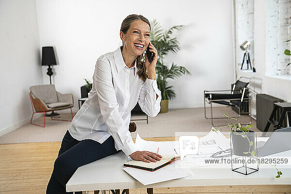 Happy mature businesswoman talking over smart phone sitting on table in office