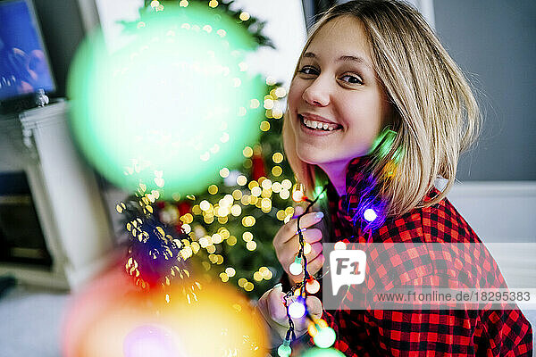 Smiling girl wearing multi colored illuminated string lights at home