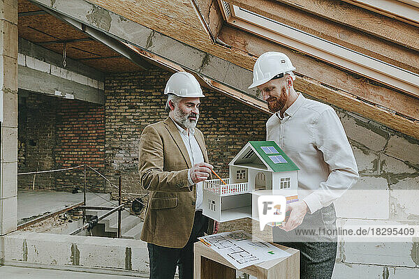 Happy architect explaining house model standing with colleague at construction site