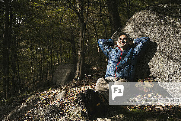 Happy mature man with hands behind head relaxing by rock in forest