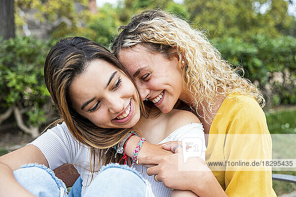 Lächelnde junge Frau mit blonden Haaren umarmt Freundin im Park